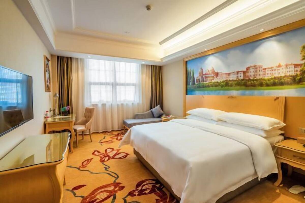 A hotel room with a bed, a desk, a chair, a TV, and a painting of a building.
