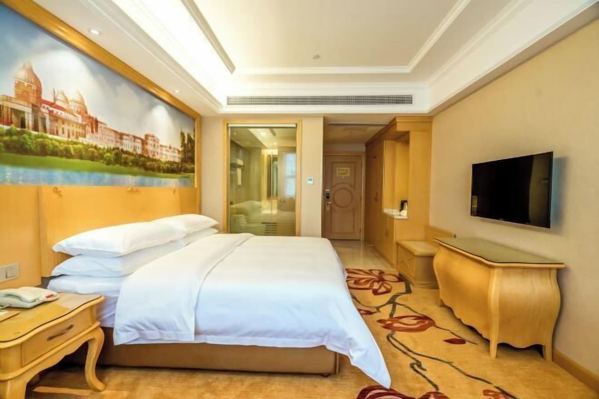 A hotel room with a bed, a TV, a painting on the wall, and a carpet.