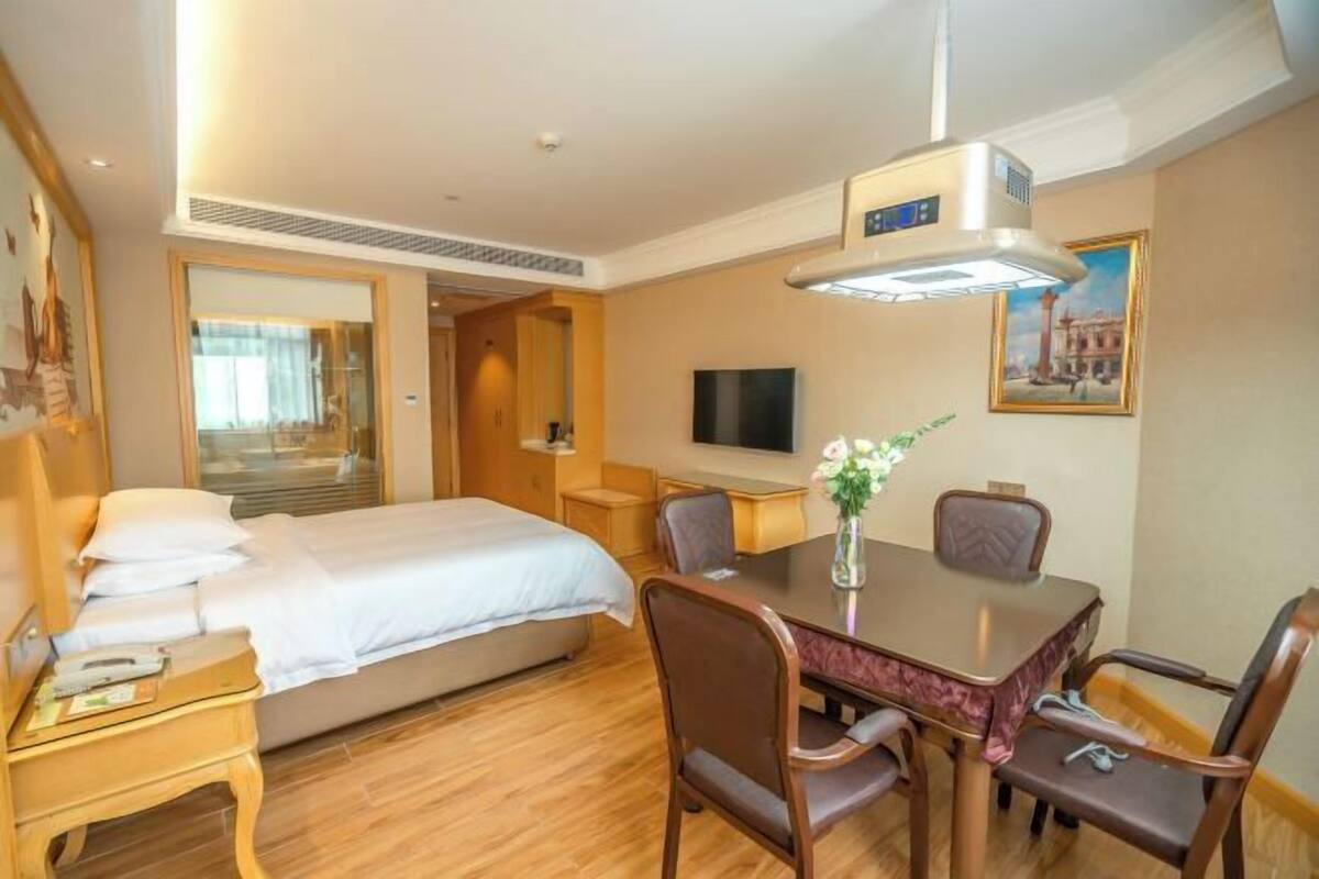 A hotel room with a bed, a dining table with chairs, a TV, and a painting on the wall.