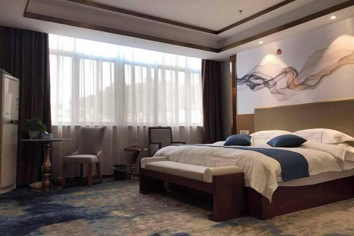 Habitación de hotel con una cama grande, un banco, un escritorio con una silla, una mesita con una planta y un ventanal con cortinas.