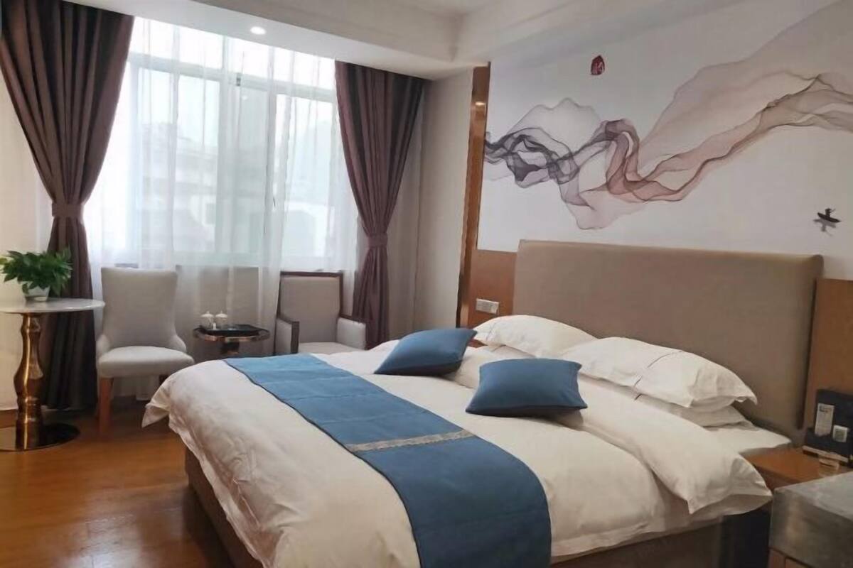 Habitación de hotel con una cama grande, un escritorio, una silla, una mesita pequeña y un amplio ventanal con cortinas.