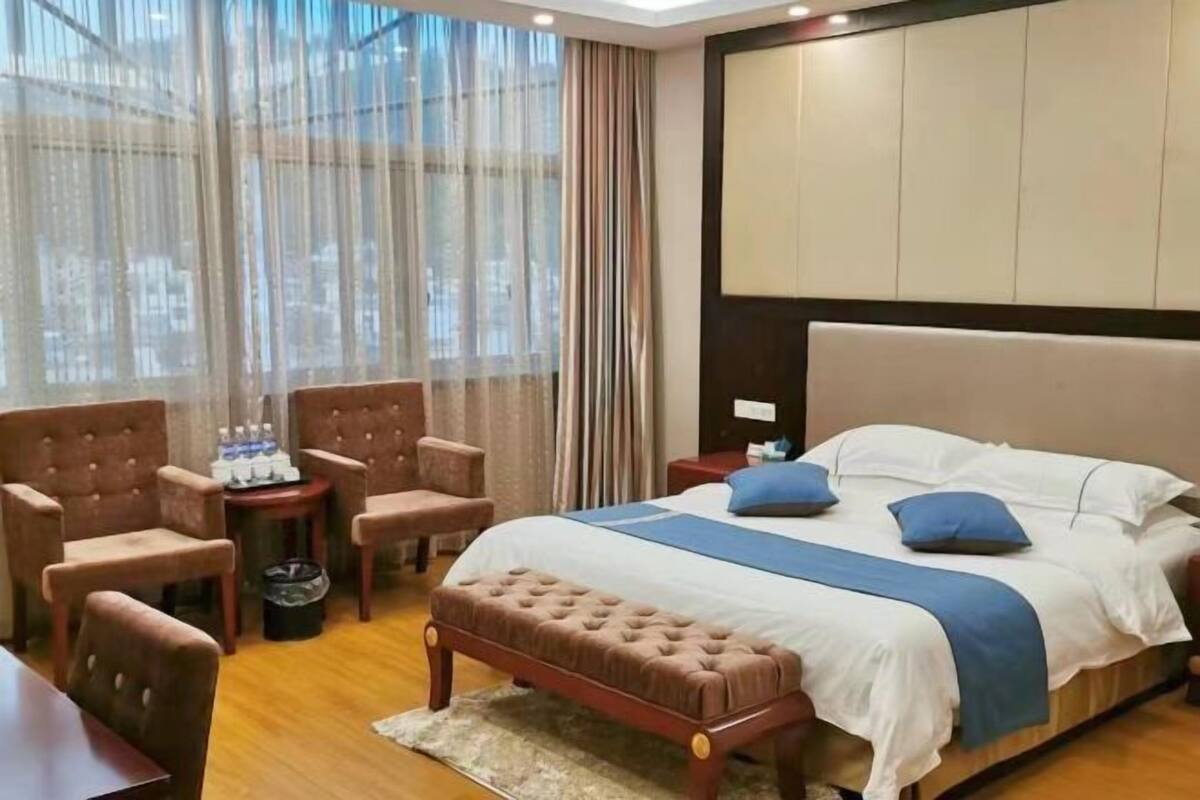 Habitación de hotel con una cama, un banco de madera, una mesita con sillas y vistas a través de cortinas traslúcidas.