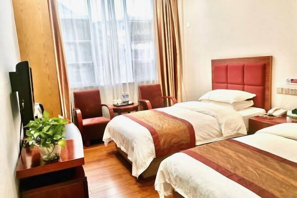 Una habitación de hotel con dos camas, un escritorio, una silla, una planta y un ventanal con cortinas.