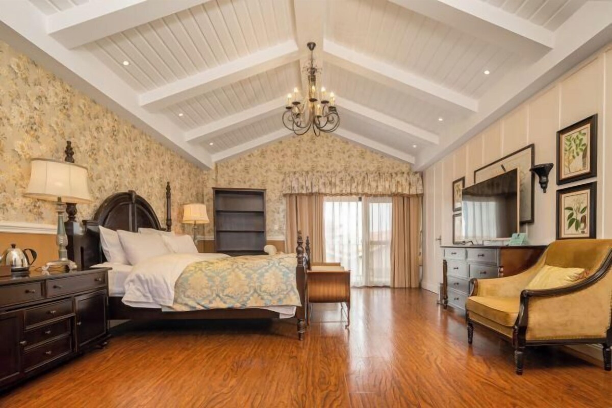 A bedroom with a wooden floor, a bed with a patterned bedspread, a chandelier, a dresser, a chair, and a large window with curtains.