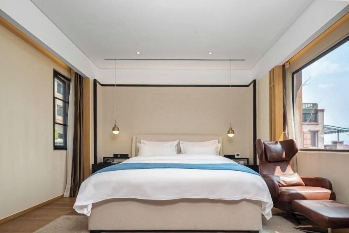 A modern hotel room with a large bed, a leather armchair, and a view of the city.