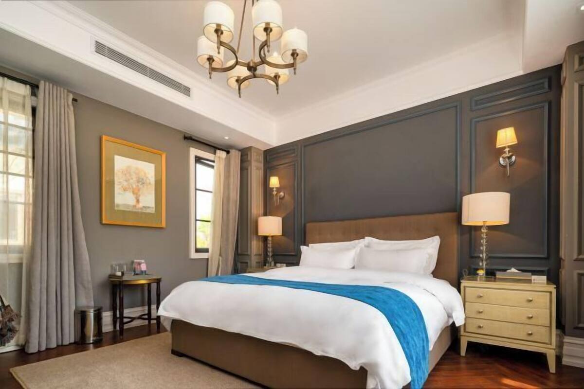 Un cuarto de hotel con una cama grande, una araña, una mesita de noche con lámpara, un cuadro en la pared y una ventana con cortinas.