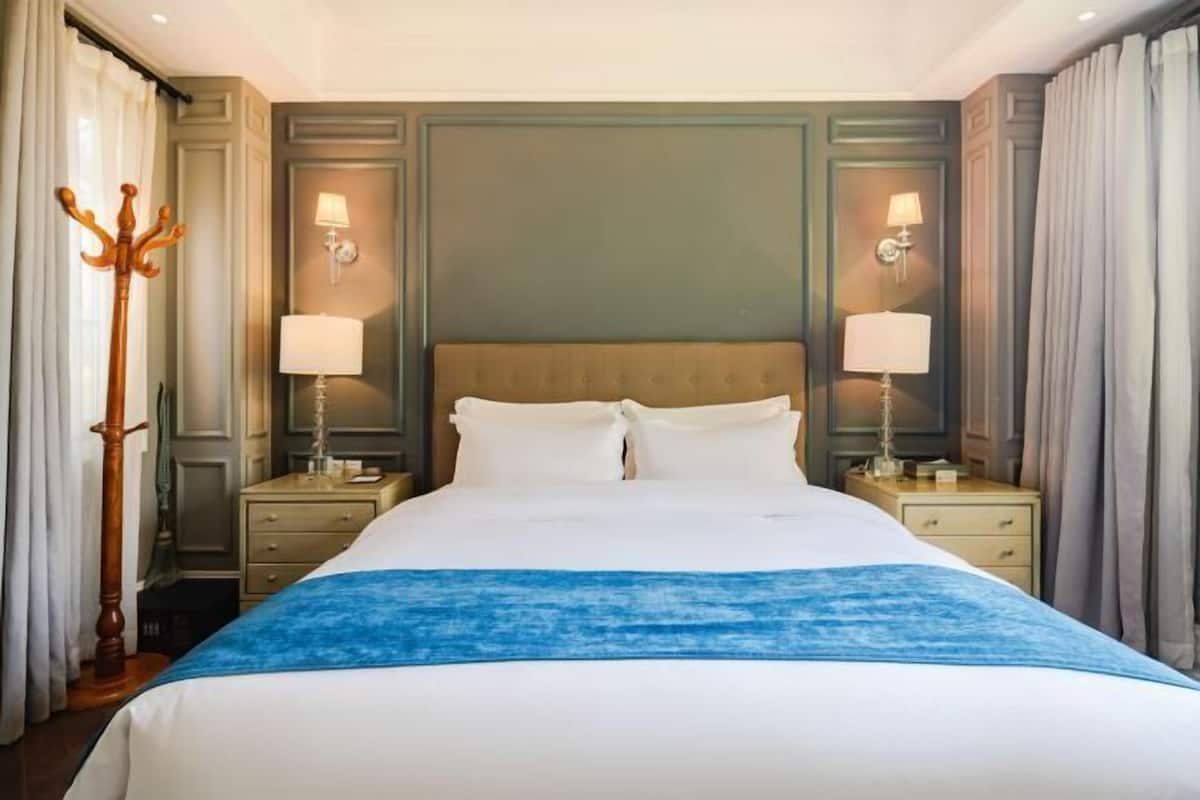 Habitación de hotel con una cama grande, dos mesitas de noche con lámparas, una lámpara de pie de madera y una pared verde decorativa.
