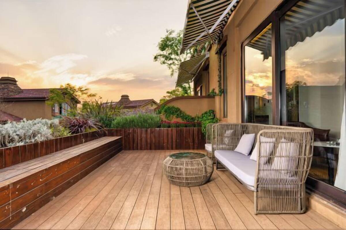 Una terraza de madera con un sofá y reposapiés de mimbre, rodeada de exuberante vegetación y vistas a casas vecinas.