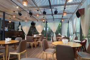 Un área de comedor interior con mesas y sillas, una zona de bar y un techo con vigas y luminarias a la vista.