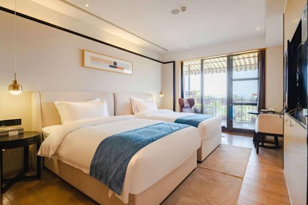 Habitación de hotel con dos camas, televisor, escritorio y balcón con vistas.