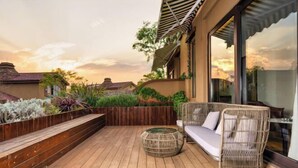 Um deck de madeira com um sofá e apoio para os pés de vime, rodeado por plantas exuberantes e um cachepot de madeira.