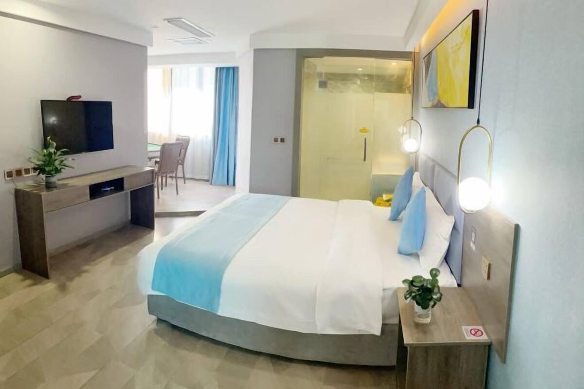 A modern hotel room with a bed, bedside table, TV, desk, and a shower area.