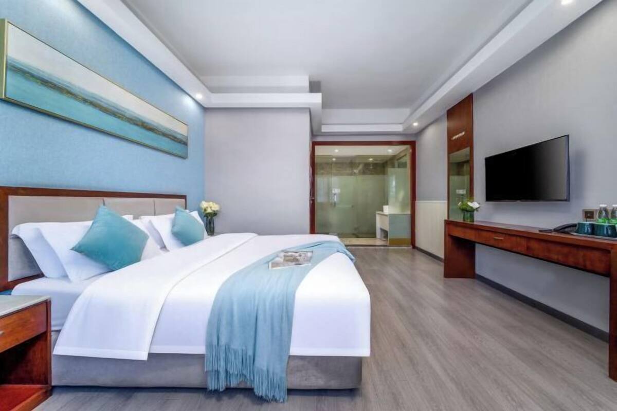 A modern hotel room with a large bed, a flat-screen TV, a wooden desk, and a glass shower area.