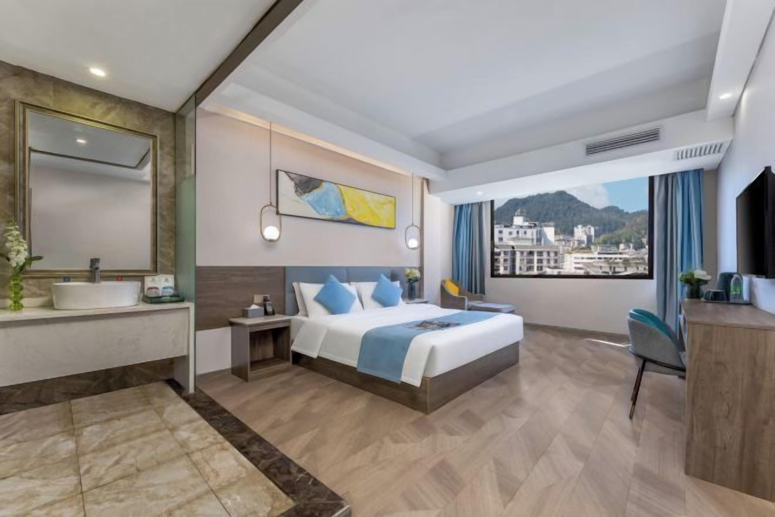 A hotel room with a bed, a desk, a bathroom with a sink, and a view of buildings through the window.