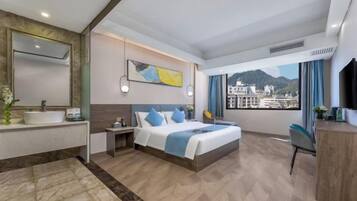 A hotel room with a bed, a desk, a bathroom with a sink, and a view of buildings through the window.