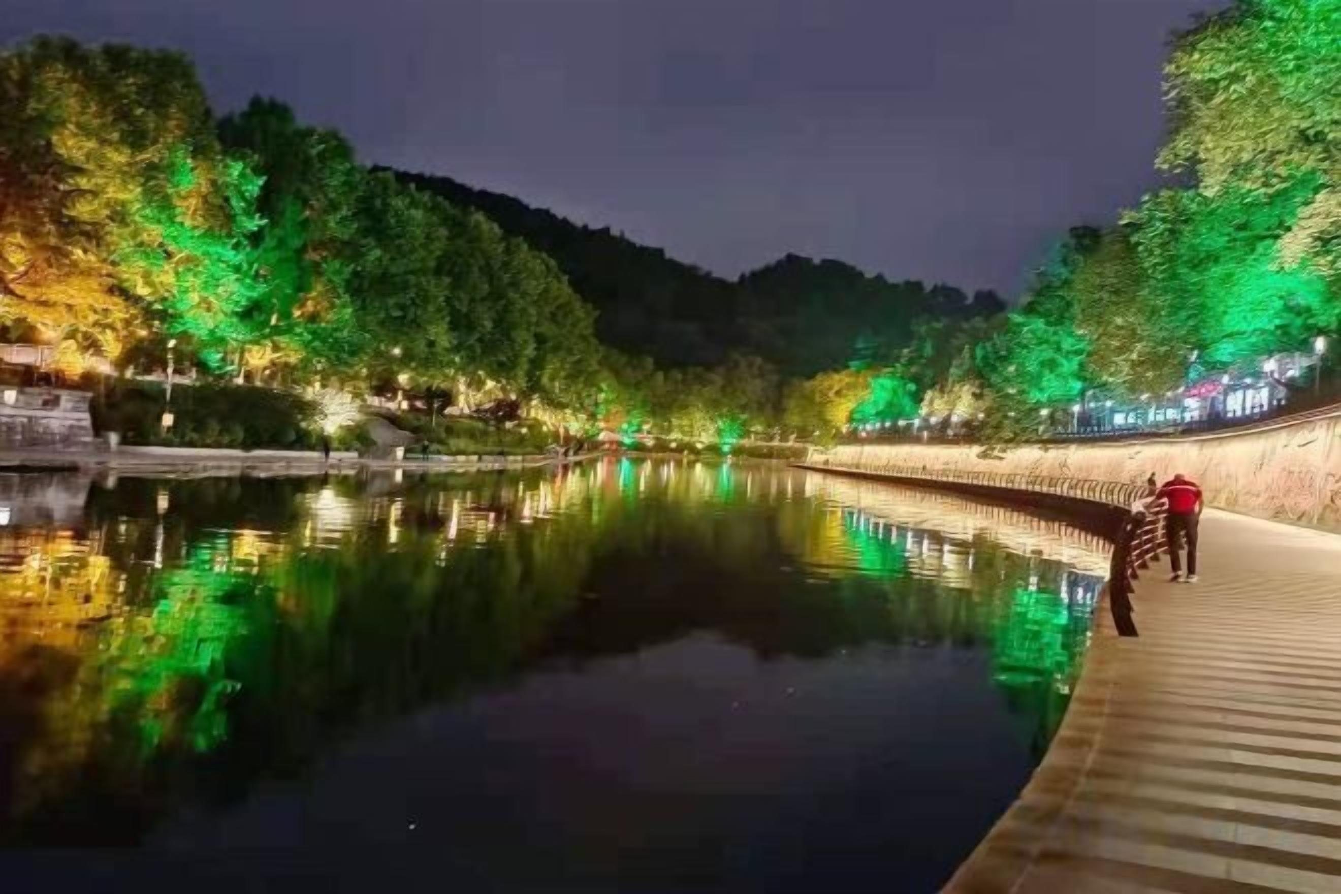 A calm river with a wooden walkway and trees on the banks, reflecting the lights.