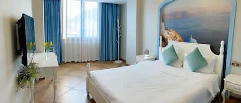A hotel room with a large bed, a TV, a desk with a vase of flowers, and a view of a coastal landscape through the window.