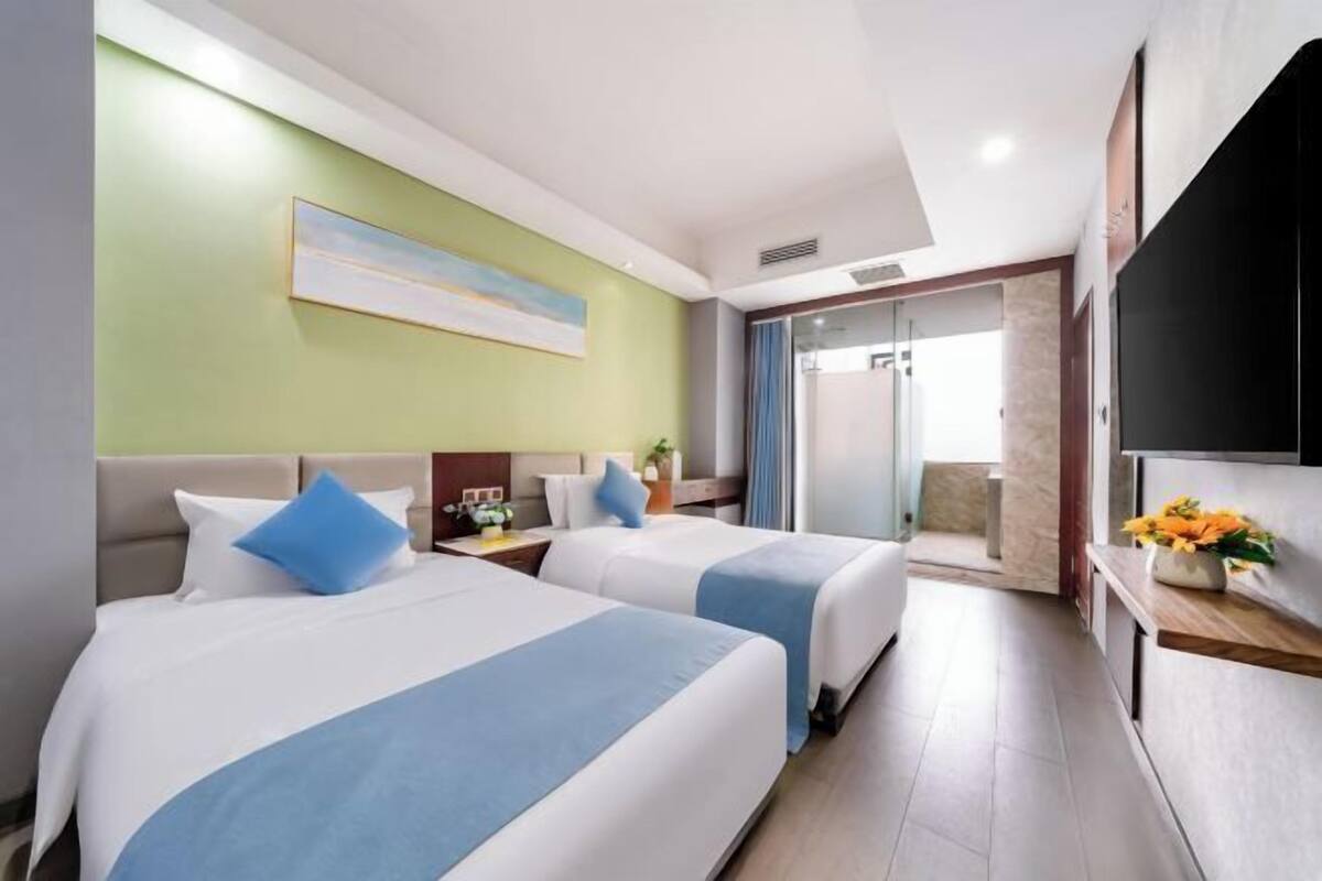 A hotel room with two beds, a television, a painting on the wall, and a bathroom visible through a glass door.