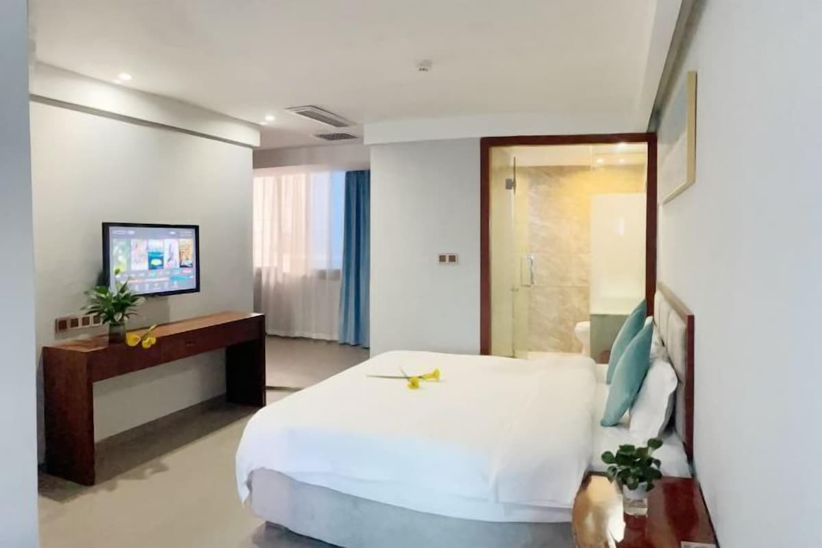 A hotel room with a bed, a TV on a stand, a desk, and a bathroom visible through a glass door.
