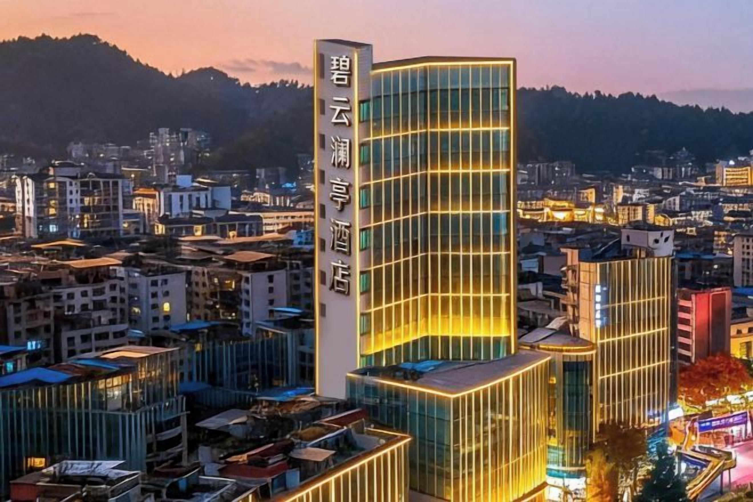 A modern hotel with a prominent vertical sign, surrounded by urban buildings and a mountainous backdrop.