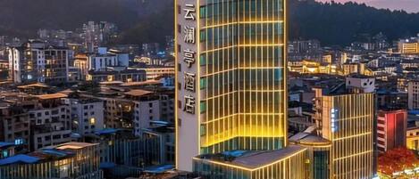 A modern hotel with a prominent vertical sign, surrounded by urban buildings and a mountainous backdrop.