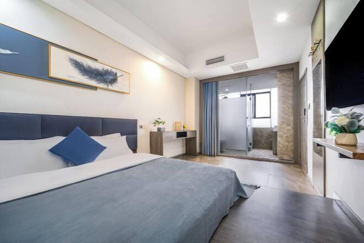 A modern hotel room with a large bed, a desk, a chair, a small table with a plant, and a sliding door leading to a bathroom.
