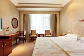 A hotel room with a large bed, a desk with a chair, a small table with two chairs, a mirror, and a window with curtains.