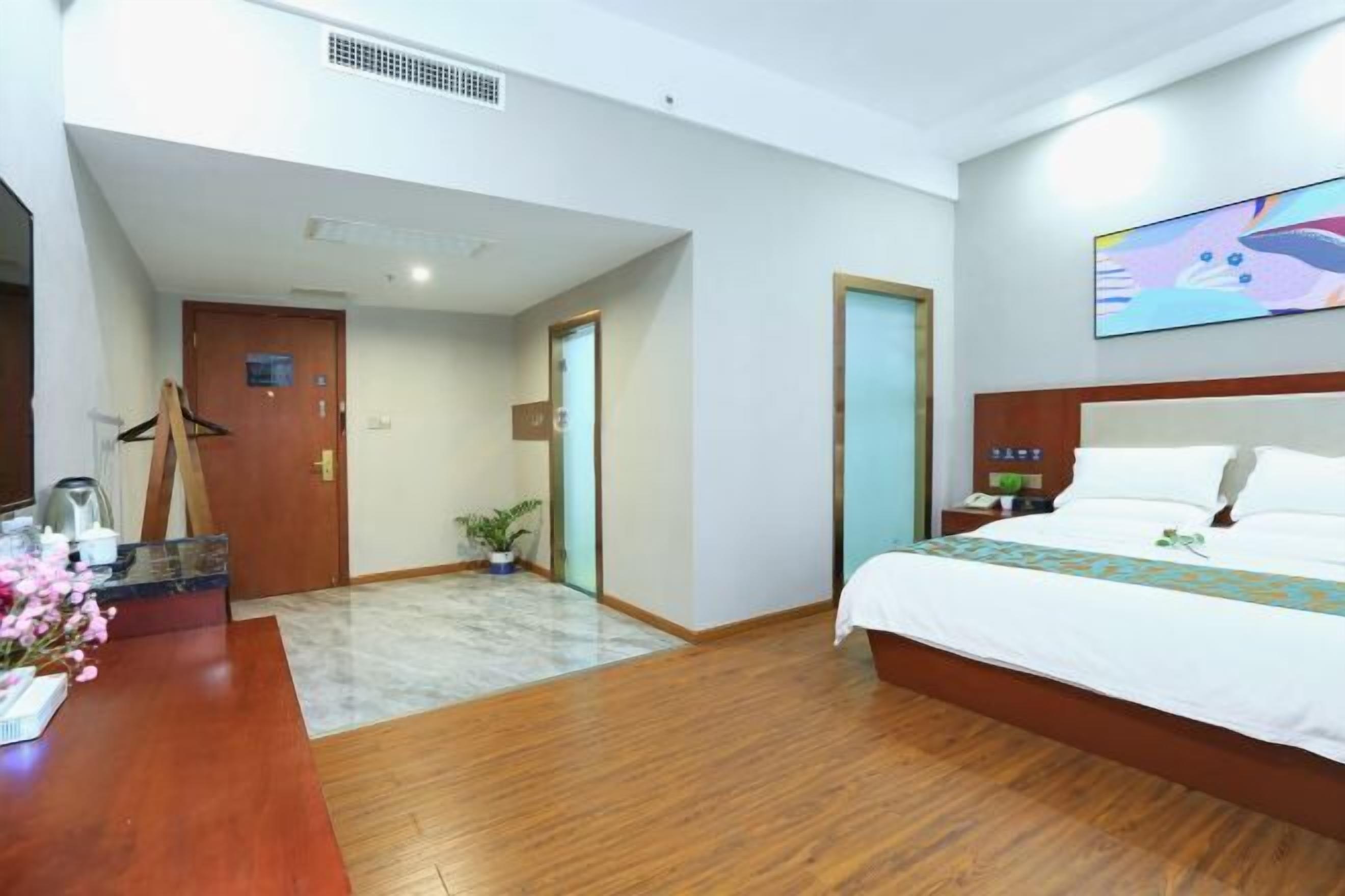 A hotel room with a wooden floor, a bed with white bedding, a desk with a coffee maker, and a painting on the wall.