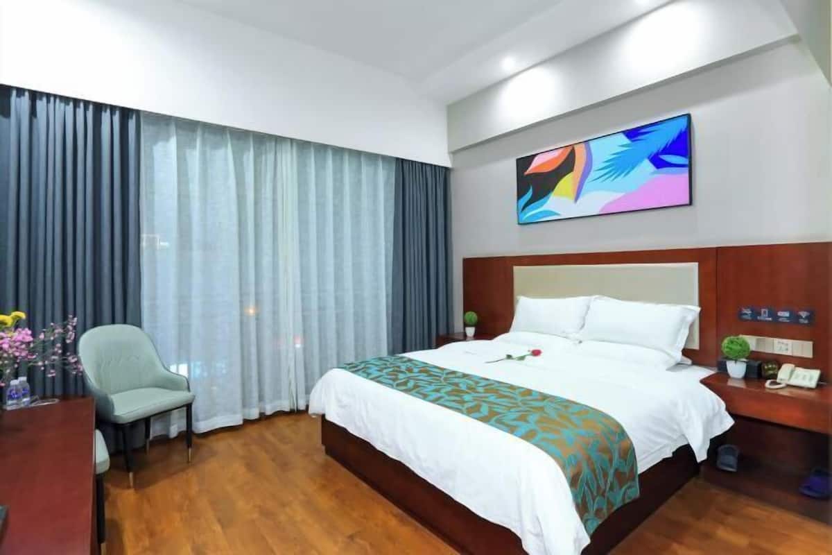 A hotel room with a bed, a desk, a chair, a nightstand, a painting, and a window with curtains.