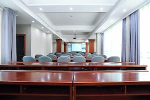Un salón de conferencias con filas de mesas y sillas de madera, ventanas amplias con cortinas y una pantalla de proyector al frente.