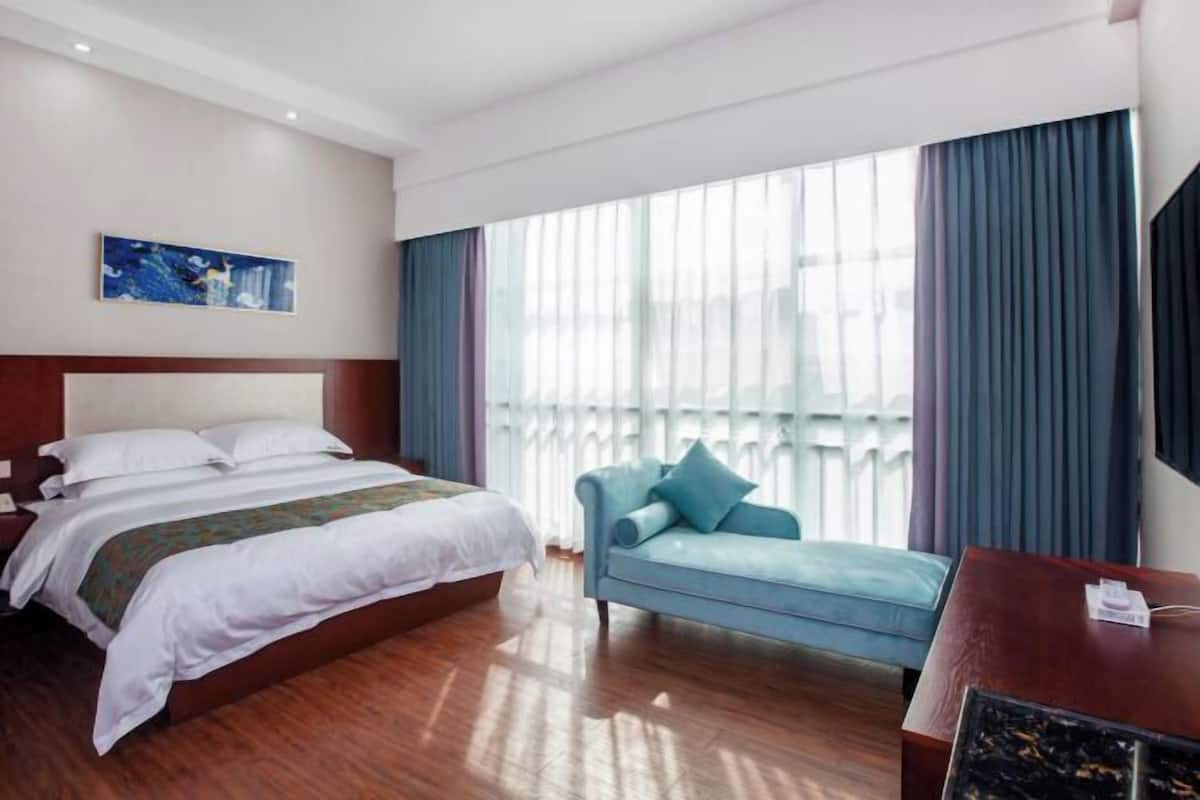 A hotel room with a bed, a blue armchair, a wooden desk, and a large window with curtains.