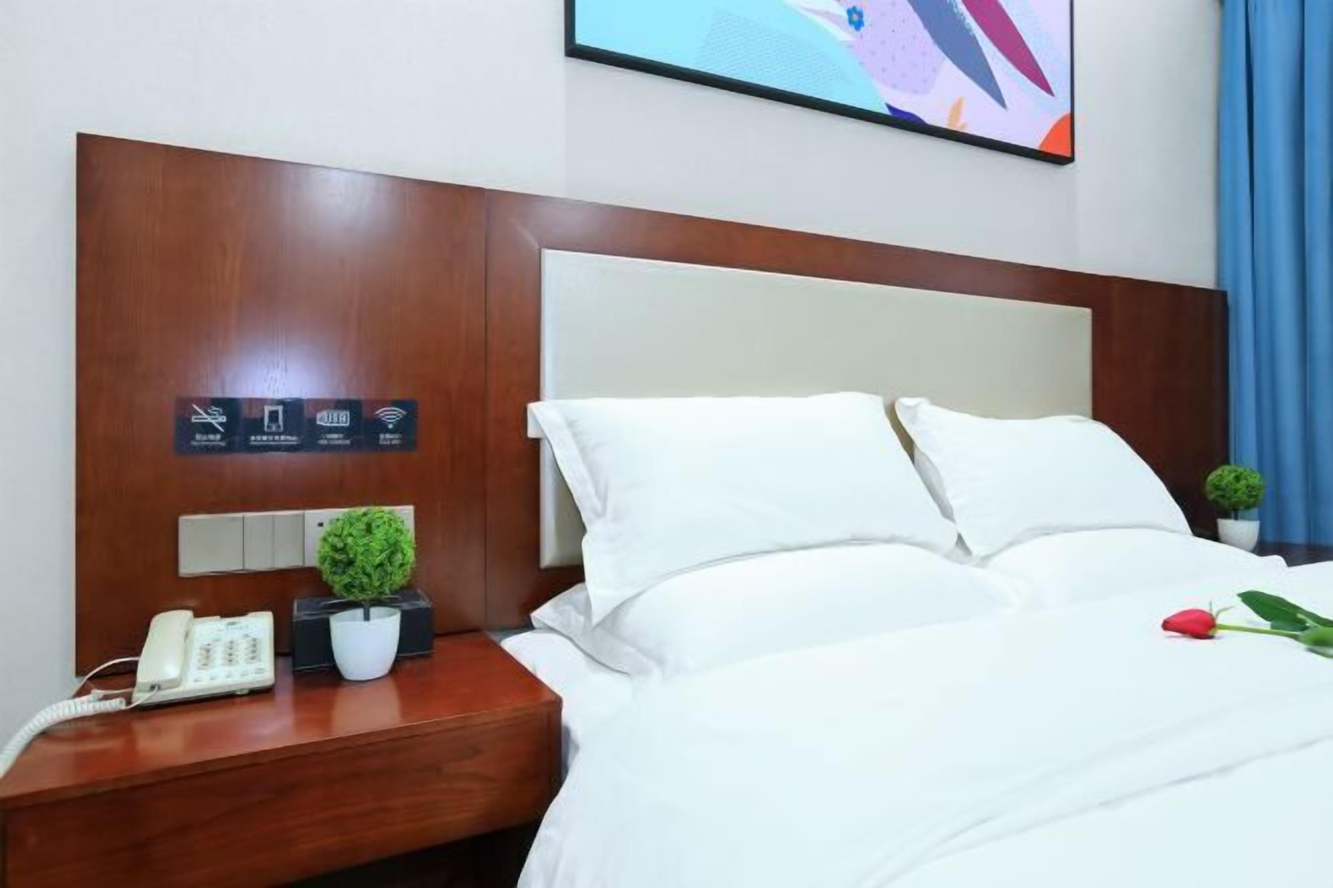 A hotel room with a wooden headboard, a bed with white linens, a bedside table with a phone, a small potted plant, and a single red rose.