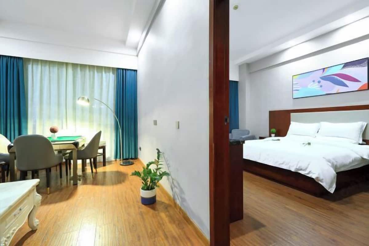 A hotel room with a bed, a desk with a chair, a potted plant, and a painting on the wall.
