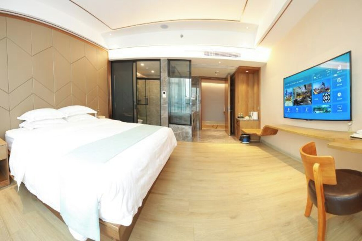A modern hotel room with a large bed, a desk, a chair, and a TV displaying a hotel app.