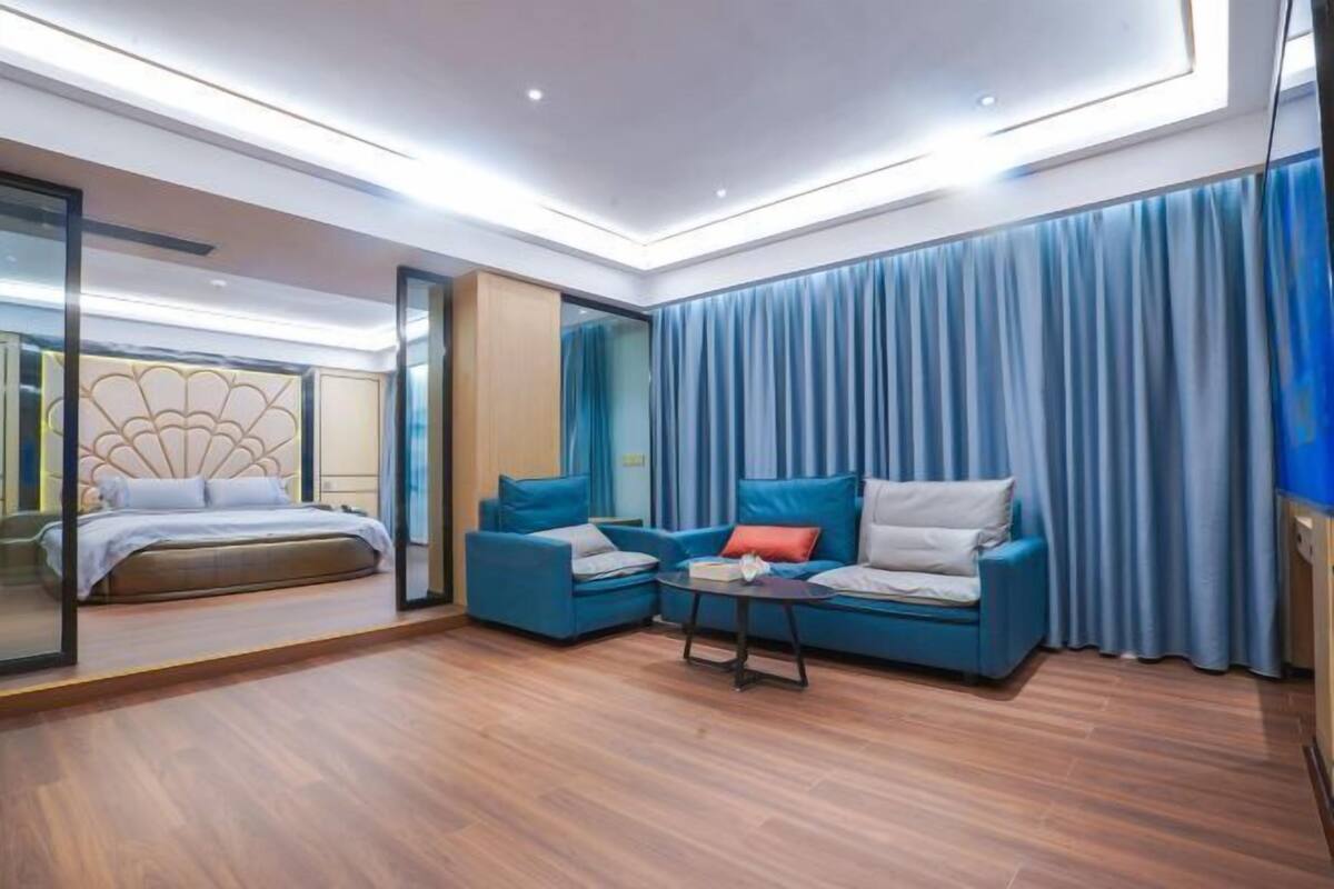 A modern hotel room with a bed, two sofas, a small table, and a large window with blue curtains.