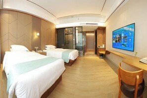 Habitación de hotel con dos camas, un escritorio, una silla, televisión y baño con puerta de vidrio.