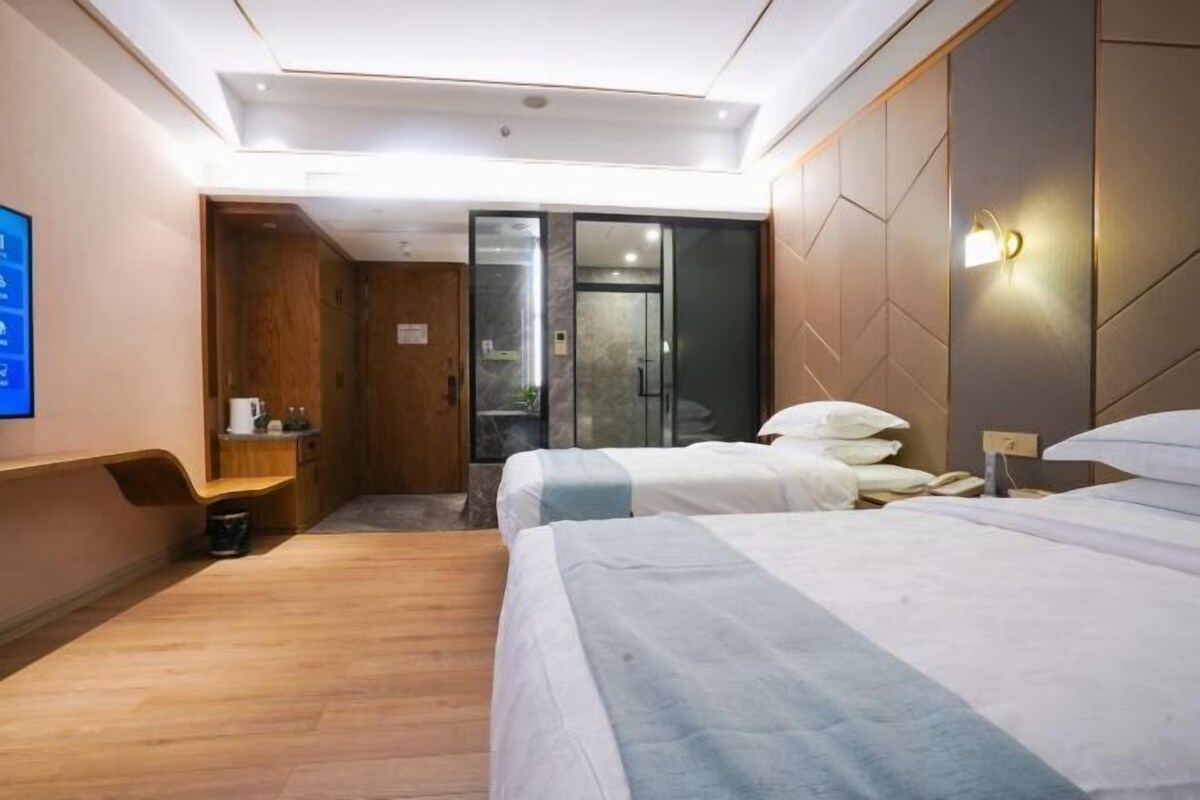 Habitación de hotel con piso de madera, cama con ropa de cama blanca y azul, mesita de noche, lámpara de pared y puerta que da a un baño.