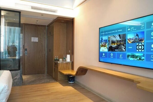 Habitación de hotel con una televisión grande de montaje en pared mostrando varias imágenes, un banco de madera, una mesita con artículos de aseo y un armario de madera.
