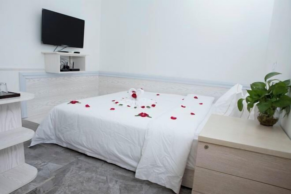 A hotel room with a bed, a TV mounted on the wall, a bedside table, a small shelf, and a potted plant.