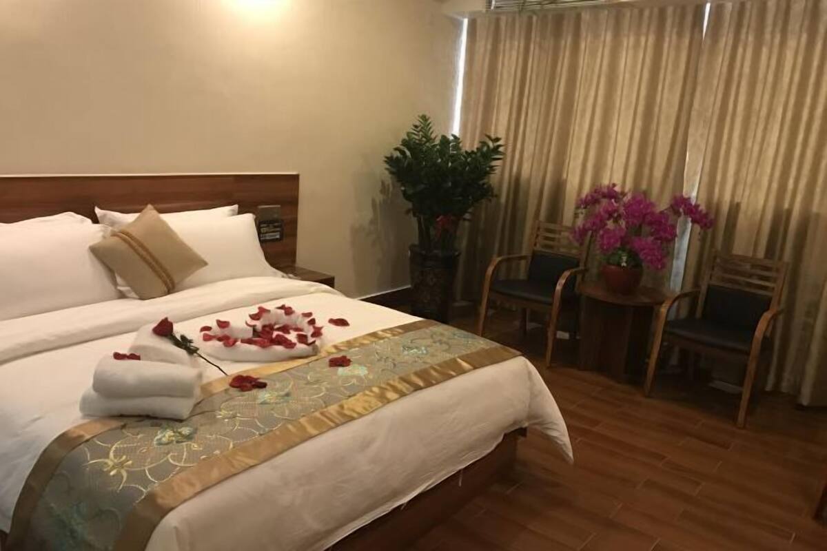 Una cama bien tendida con una colcha floral, una mesita de noche con una lámpara, una silla, una pequeña mesa con una planta en maceta y una ventana con cortinas.