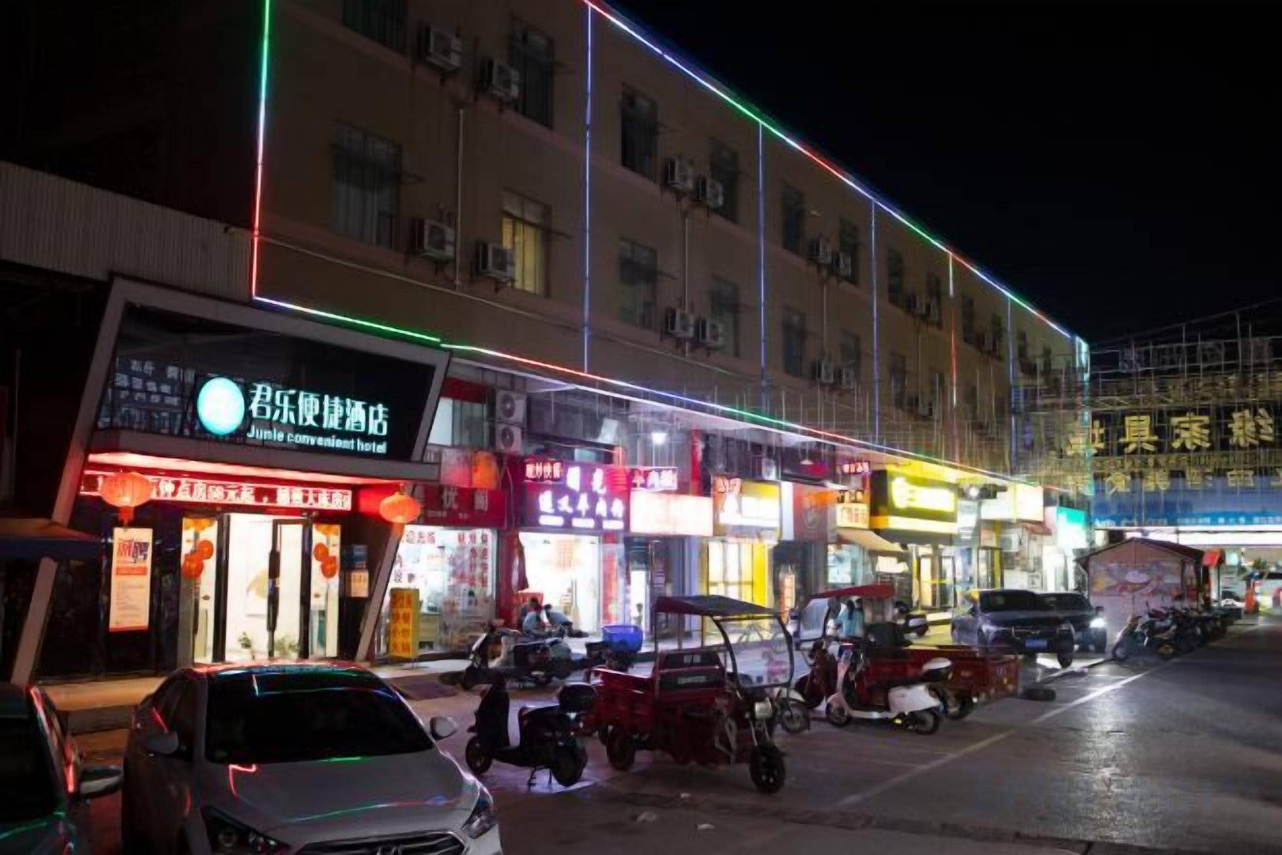 A commercial street at night with illuminated storefronts and parked vehicles.