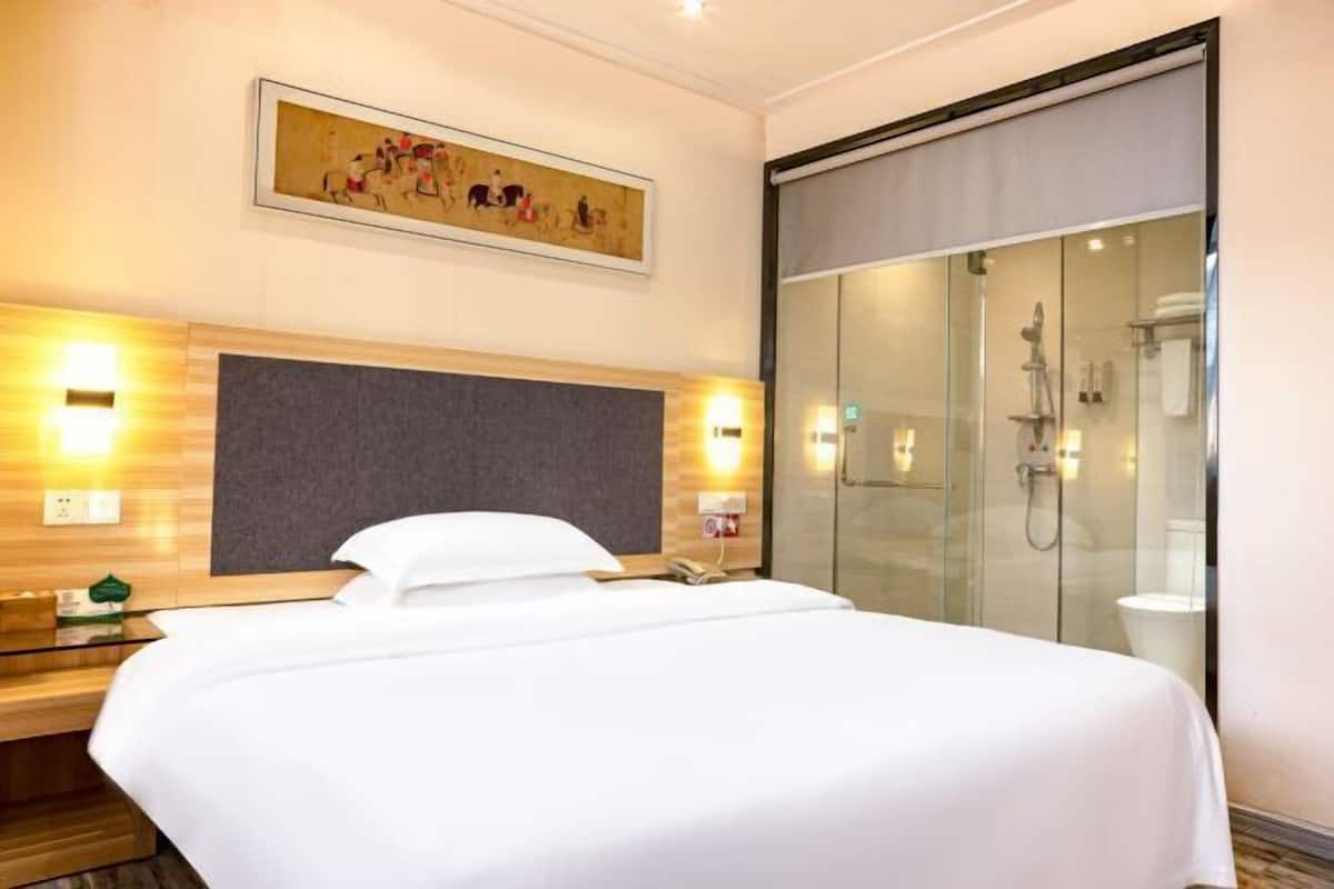 A hotel room with a bed, a nightstand, a wall-mounted phone, a painting, and a glass-enclosed shower area.