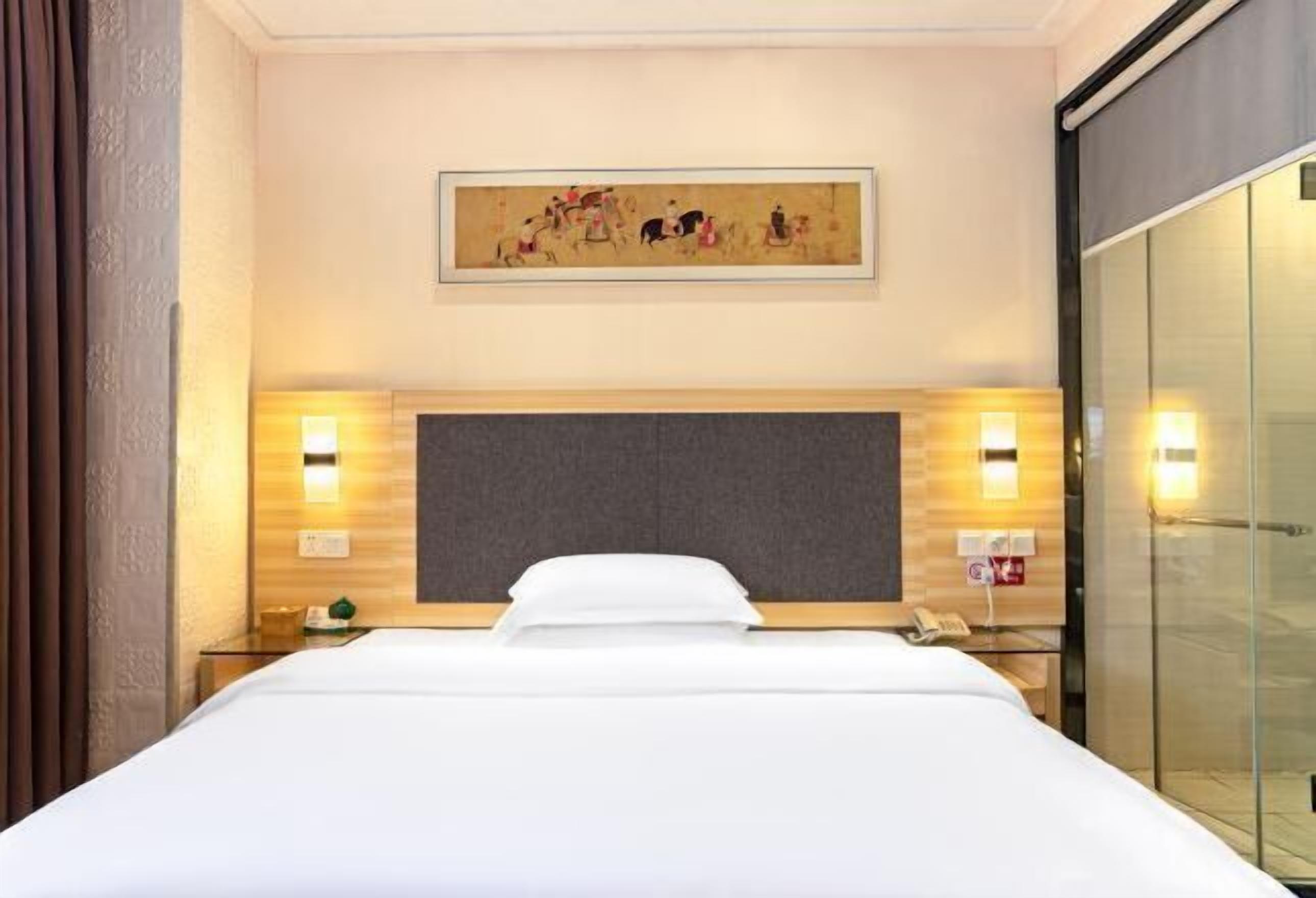 A hotel room with a large bed, a headboard, bedside tables with lamps, a painting on the wall, and a sliding glass door.
