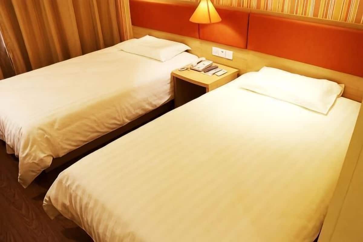 Two single beds with white linens, a wooden nightstand with a lamp, and a headboard with a light fixture.