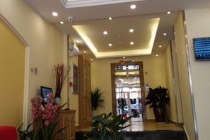 Um saguão de hotel com balcão de recepção, plantas em vasos e uma escada que leva aos andares superiores.