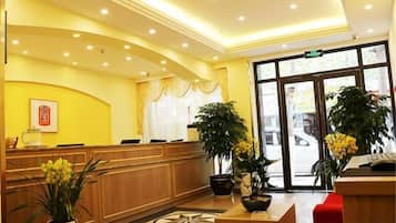 A hotel reception area with a wooden reception desk, a large potted plant, and a tiled floor.