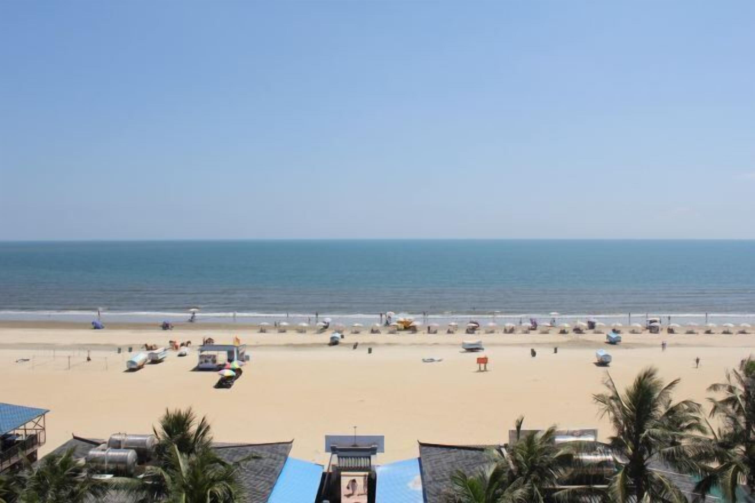 A beach view with a clear sky, palm trees, and a sandy beach with umbrellas and lounge chairs.