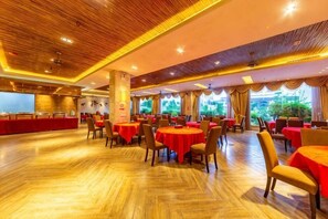 Un vaste espace de restauration intérieur, doté de plusieurs tables et chaises rondes, d’un plafond en bois et de grandes fenêtres agrémentées de rideaux.
