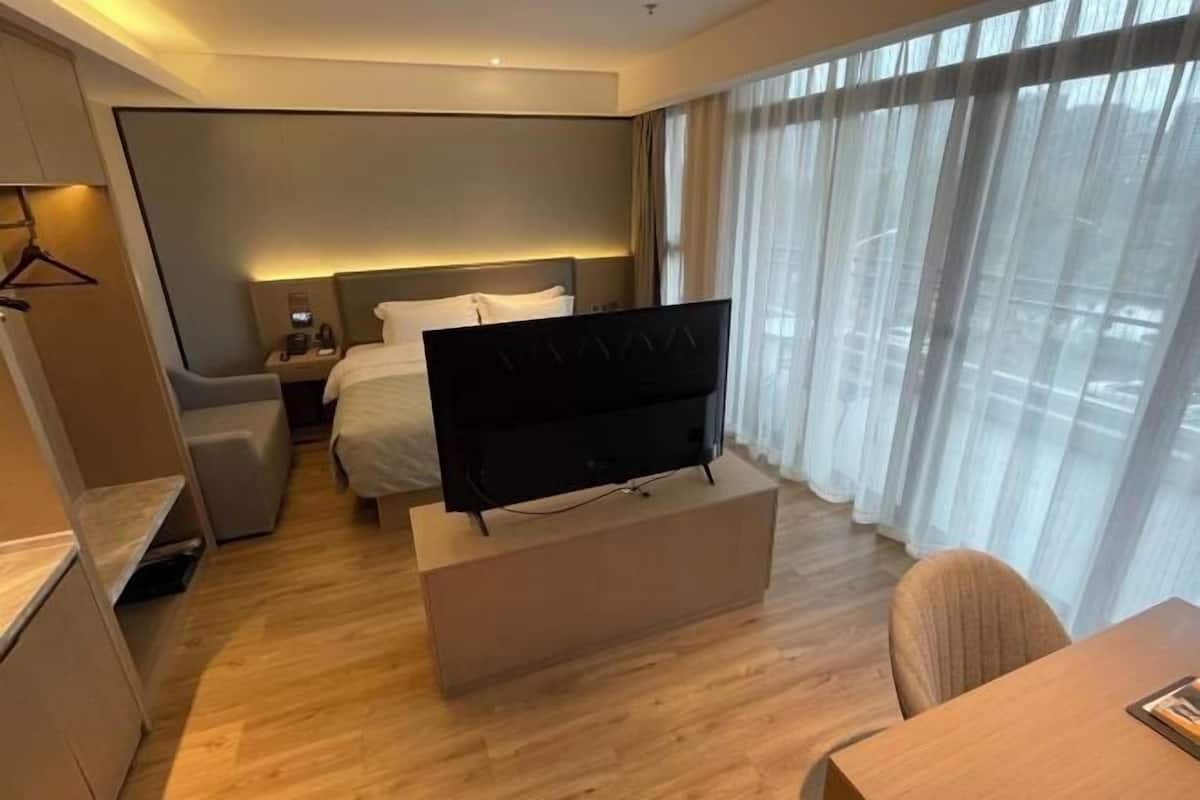 Ein Hotelzimmer mit Bett, Nachttisch, Fernseher auf einem Standfuß und einem Fenster mit Vorhängen.