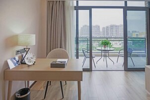 Habitación de hotel moderna con un escritorio de madera, una silla, una pequeña mesa con una planta y vistas a edificios altos a través de amplios ventanales.